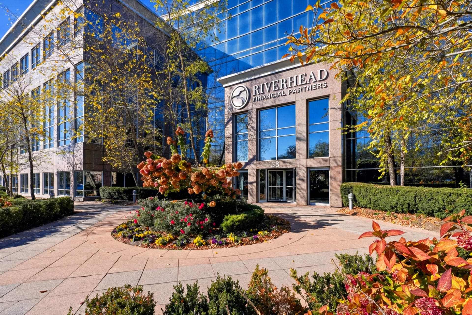 Riverhead Financial Partners' office building in Melville, New York — a glass-and-granite entrance framed by autumn trees.